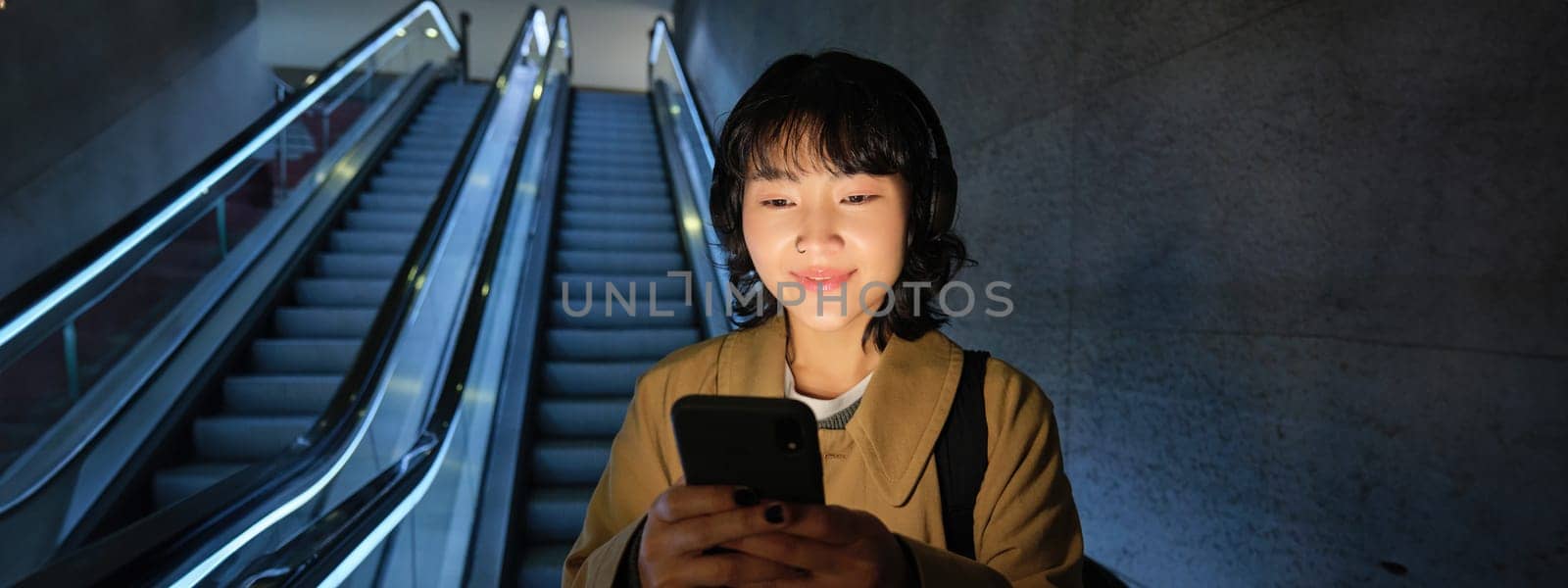 Happy smiling young woman, standing on escalator, going down, holding smartphone in both hands, chatting on mobile phone app by Benzoix