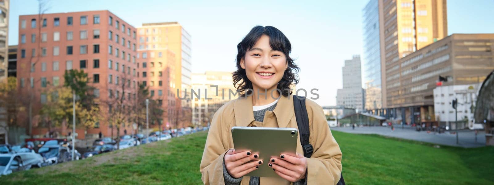 Happy asian girl stands on street, university student walks with digital tablet in hands and smiles, stands in city centre.