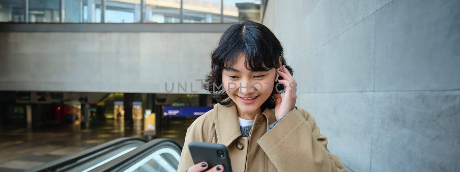 Portrait of cute korean girl in trench coat, going up escalator, holds mobile phone and smiles pleased, watches video in wireless headphones by Benzoix