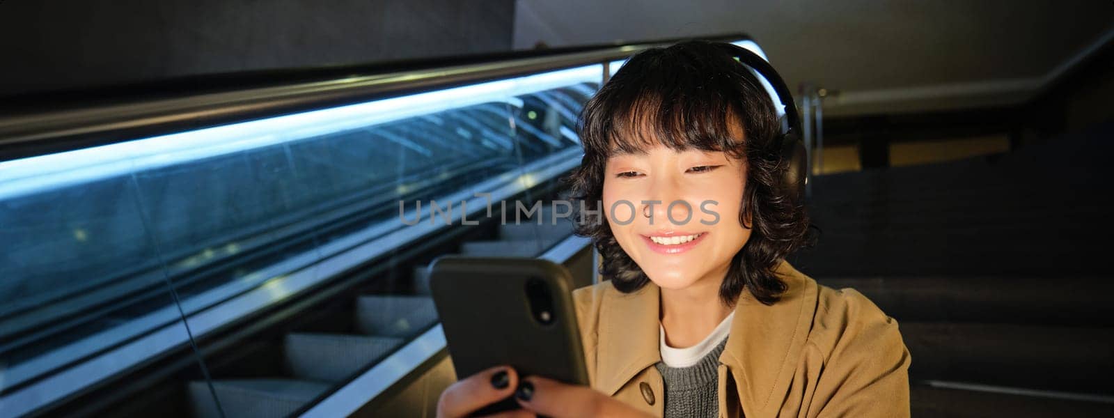 Close up portrait of smiling asian girl student, listens music in headphones and looks at mobile phone, uses smartphone, sits on staircase in public place by Benzoix