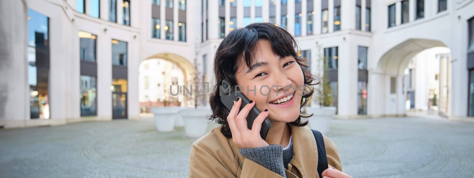 Portrait of smiling asian girl talks on mobile phone, speaks with friend on smartphone, walks in city centre, stands on street and laughs by Benzoix