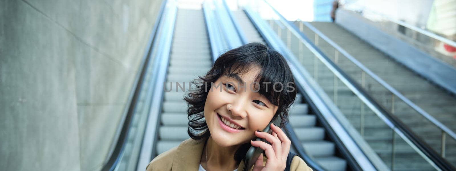 Close up portrait of smiling brunette girl, korean woman goes down escalator, makes phone call, talks to someone on telephone.