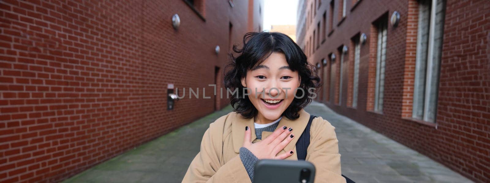 Portrait of amazed girl, looks at smartphone screen and gasps from happiness, reads good news on text message, being impressed by smth on mobile phone by Benzoix
