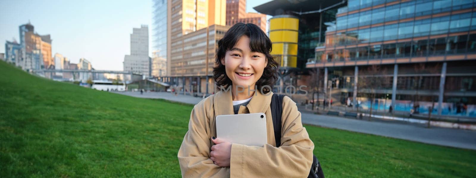 Happy asian girl stands on street, university student walks with digital tablet in hands and smiles, stands in city centre.