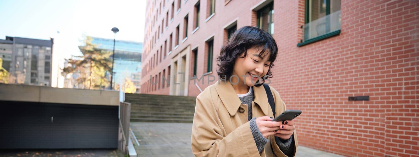 Stylish korean hipster girl, walks in city centre, sends text message, uses location map application on smartphone while walking down the street, wears trench coat by Benzoix