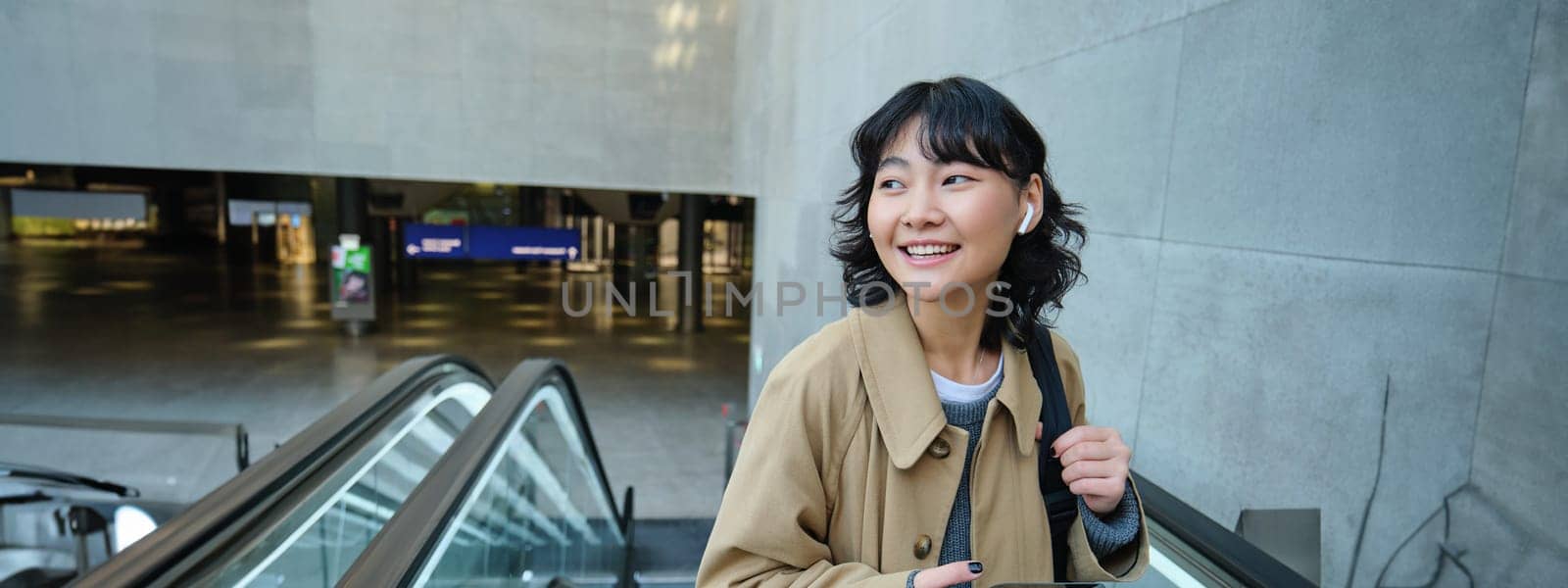 Portrait of brunette girl in wireless headphones, listens music, uses smartphone, goes on escalator by Benzoix