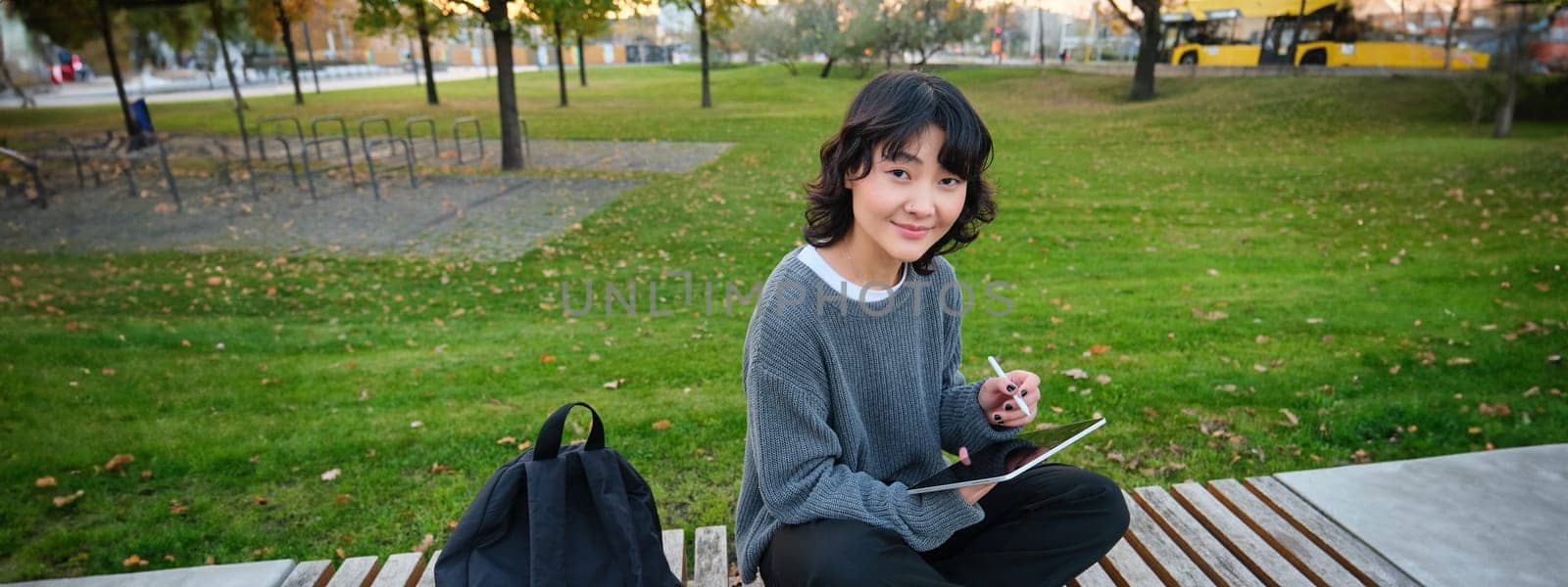 Young asian girl with graphic pencil and tablet, sits in park on bench, draws scatches, does her homework outdoors.