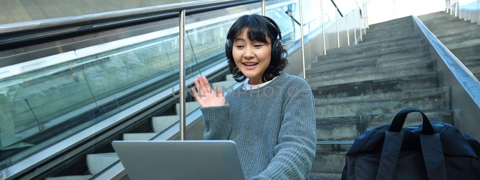 Smiling young student, wears headphones, sits on street stairs and waves hand at her laptop, connects to video chat, conference or training session on remote by Benzoix