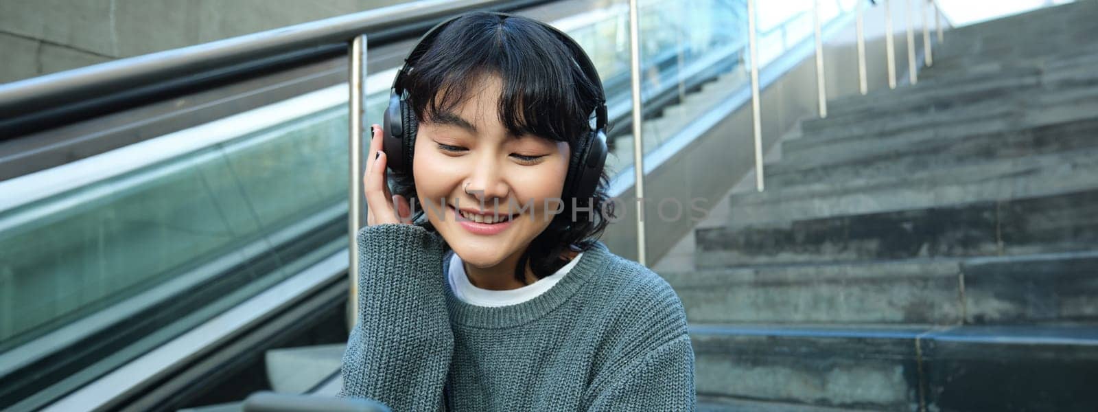 Portrait of smiling korean girl in headphones, uses smartphone and sits on stairs in mall, watches video on mobile phone by Benzoix