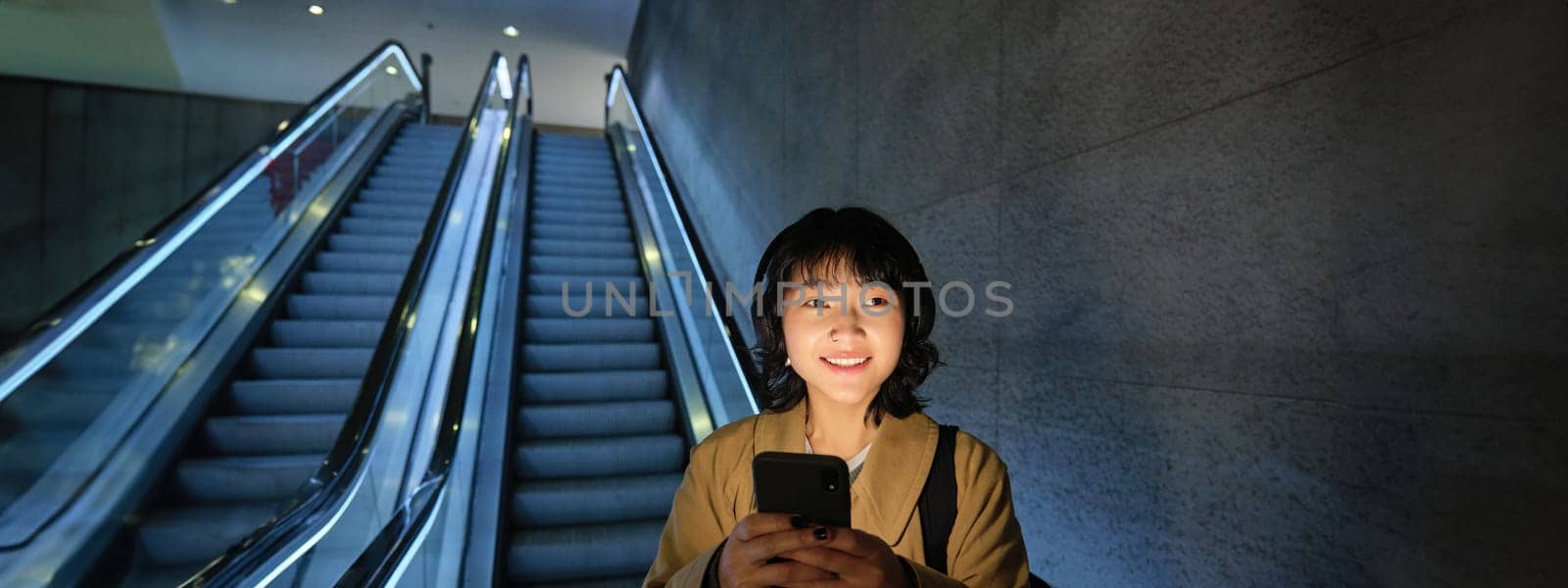 Cute young woman going down the escalator to the tube, using subway metro to commute to work or university, standing with smartphone.