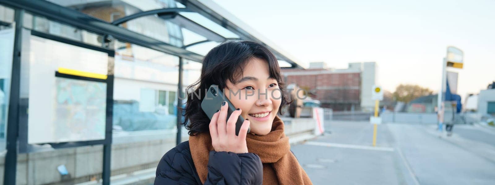Smilling Korean girl talking on mobile phone, standing on bus stop, using smartphone, posing on road in winter, wrapped in scarf, wearing black jacket.