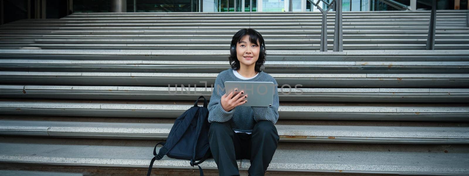 Young woman draws on her tablet, listens to music in headphones. Asian girl sits on stairs and does graphic design project, sits on street stairs by Benzoix