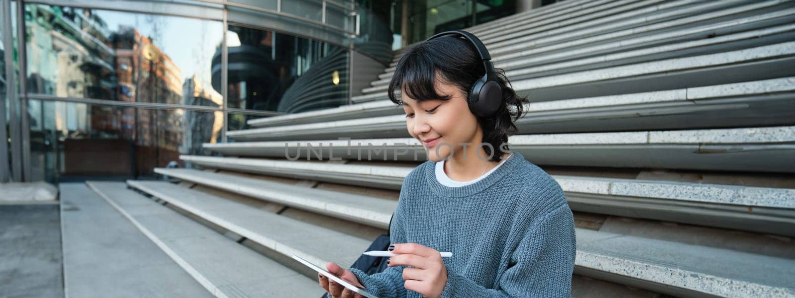 Young smiling digital artists works on her project using tablet and graphic pen, listens music in headphones and sits on stairs in public space, works on project.