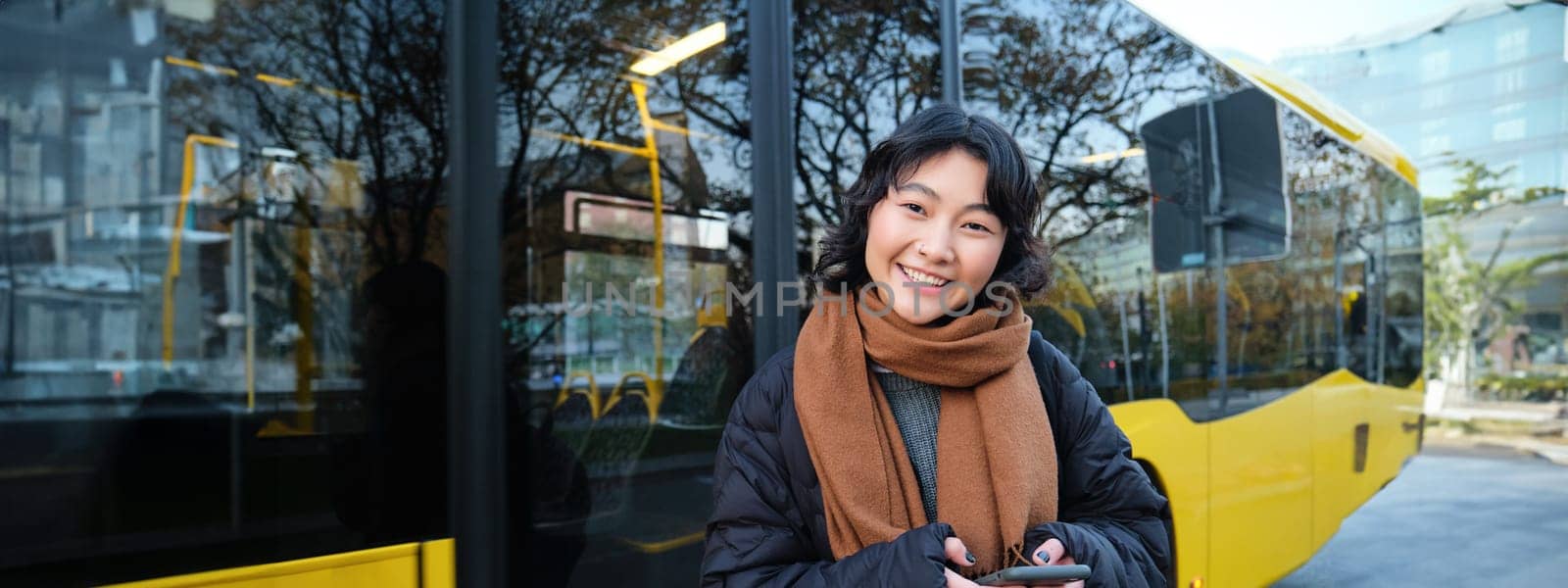 Portrait of korean girl buying ticket for public transport online, using mobile application on bus stop, wearing winter clothes.
