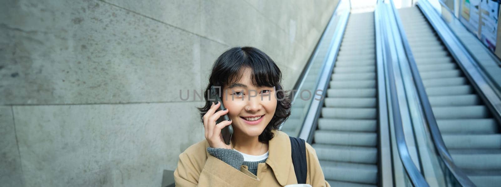 Portrait of smiling korean girl commutes, goes somewhere in city, drinks coffee to go and uses smartphone, stands on escalator by Benzoix