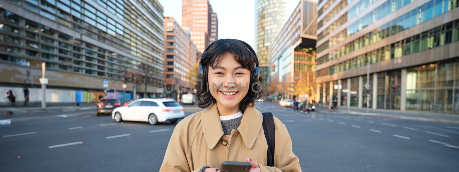 Portrait of young asian girl, student walks in city, listens music in headphones and uses mobile phone on streets by Benzoix