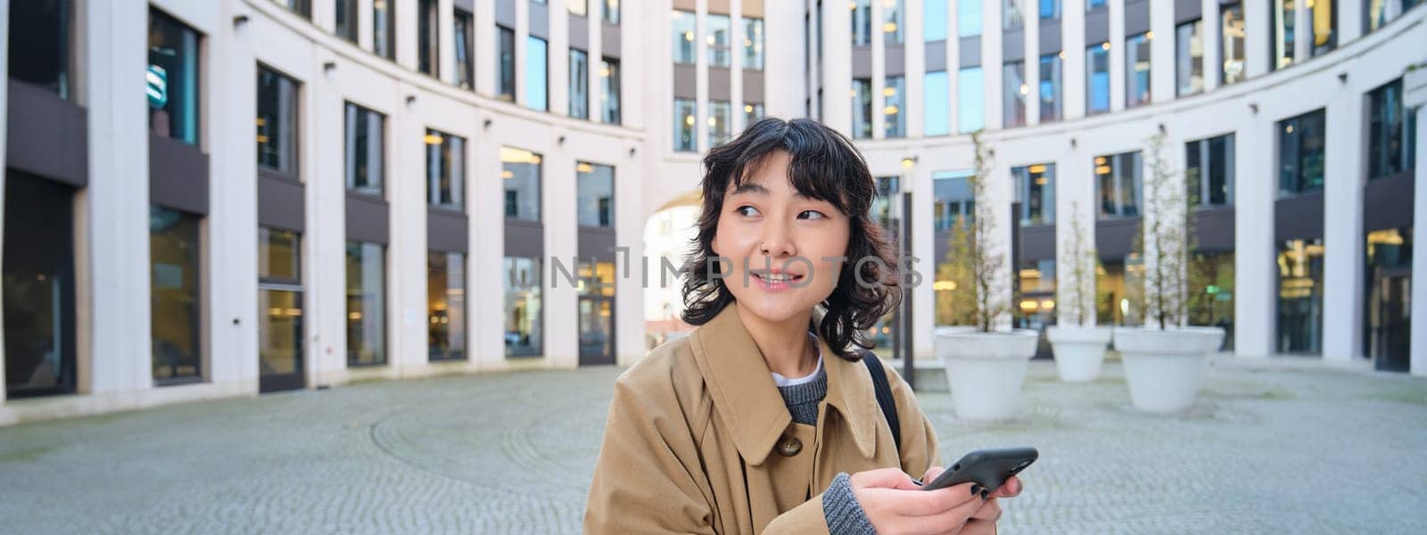 Stylish korean girl in headphones, listens music and uses mobile phone, stands in city centre, waits for someone on street and writes text message on her smartphone by Benzoix