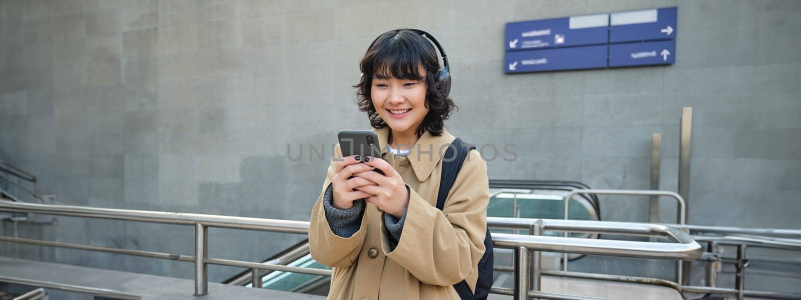 Portrait of stylish asian hipster girl, listens music in headphones, looks at her phone, tourist looking at map for a way to sightseeing, drinking coffee to go by Benzoix