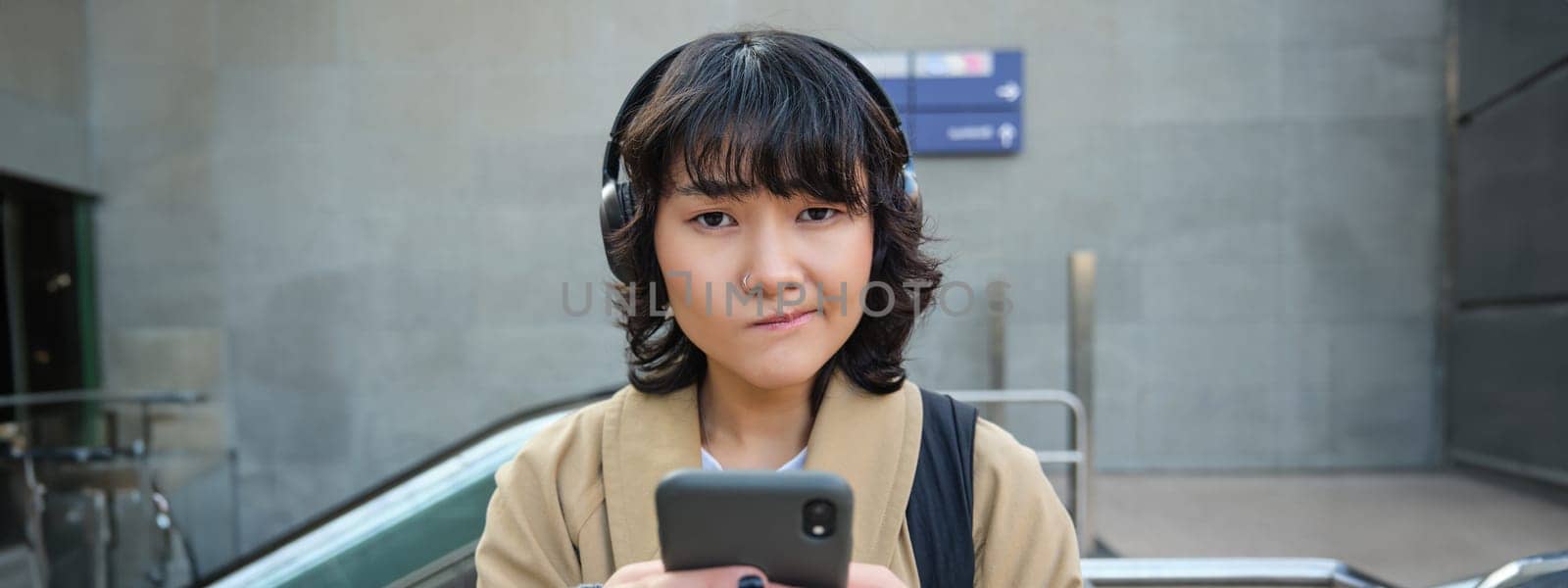 Close up portrait of puzzled, complicated korean girl in headphones, frowns and looks unsure, stands with smartphone and looks at camera while shrugs on street by Benzoix