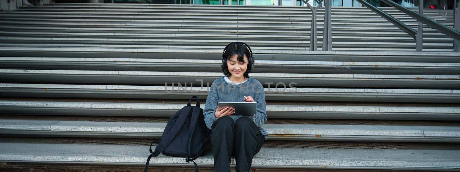 Young smiling digital artists works on her project using tablet and graphic pen, listens music in headphones and sits on stairs in public space, works on project.