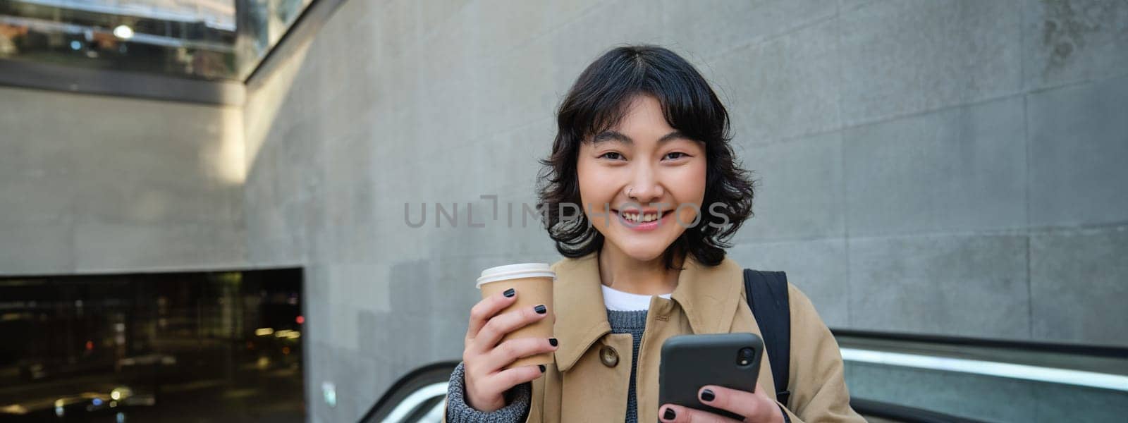 Portrait of hipster girl on escalator, drinking coffee and looking at smartphone, going to work, commute in city, smiling at camera by Benzoix