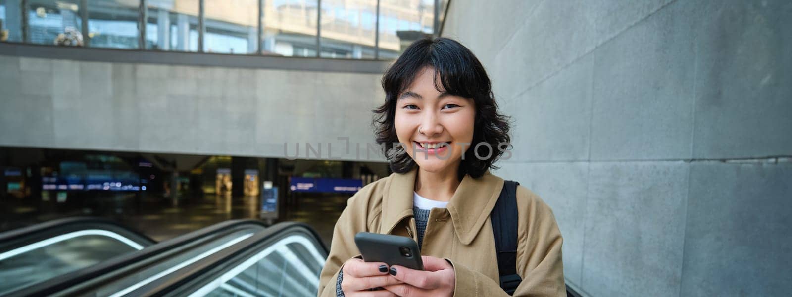 Young brunette woman commutes, goes somewhere in city, stands on escalator and uses mobile phone, holds smartphone and smiles.
