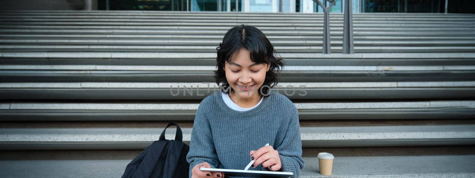 Smiling girl, graphic designer, using digital tablet and pen tool to draw, does home assignment for university, sits on stairs.
