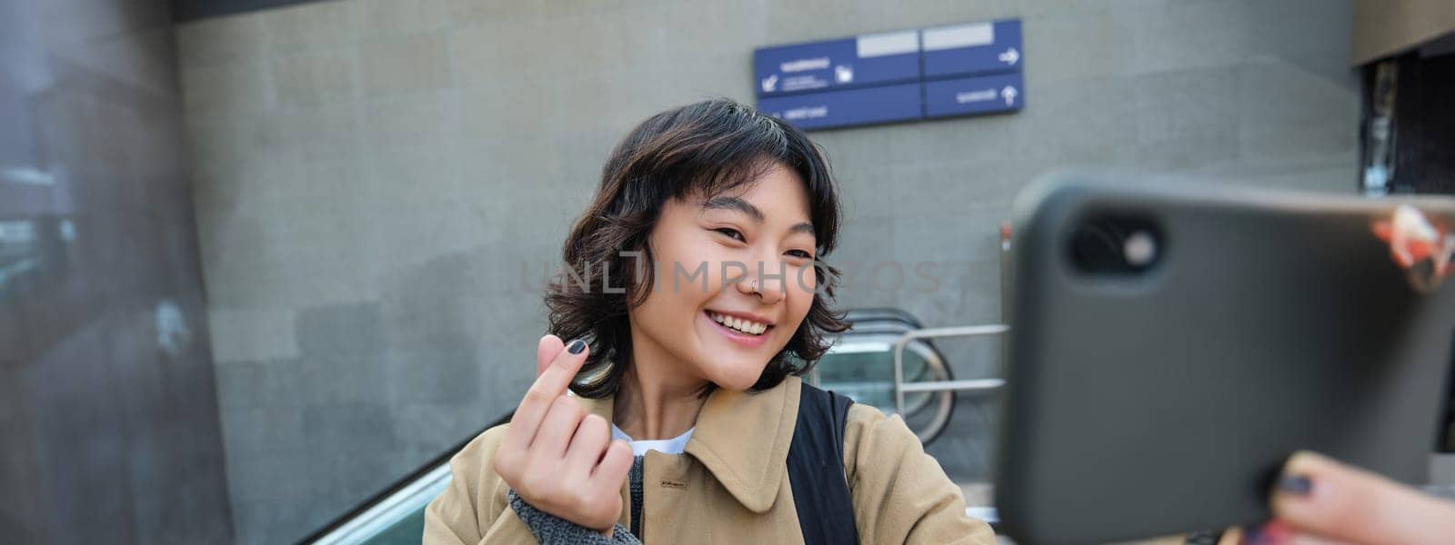 Portrait of stylish korean girl, student takes selfie on street, holds smartphone and poses on its camera, makes video on tourist trip, smiles with joy by Benzoix