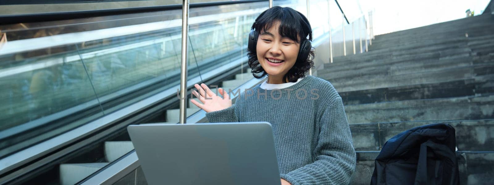 Smiling young student, wears headphones, sits on street stairs and waves hand at her laptop, connects to video chat, conference or training session on remote by Benzoix