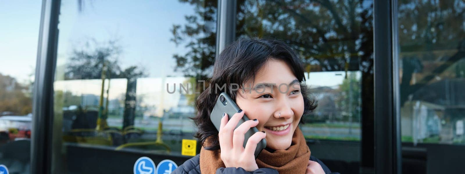 Cellular technology and people concept. Stylish asian girl talks on mobile phone, makes a telephone call, stands near bus stop and has conversation by Benzoix