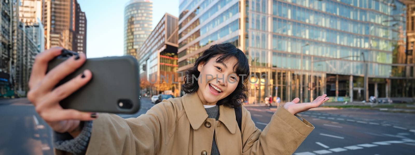 Upbeat asian girl takes selfie with smartphone in city centre, showing something behind her with smiling face. Tourist goes sightseeing.