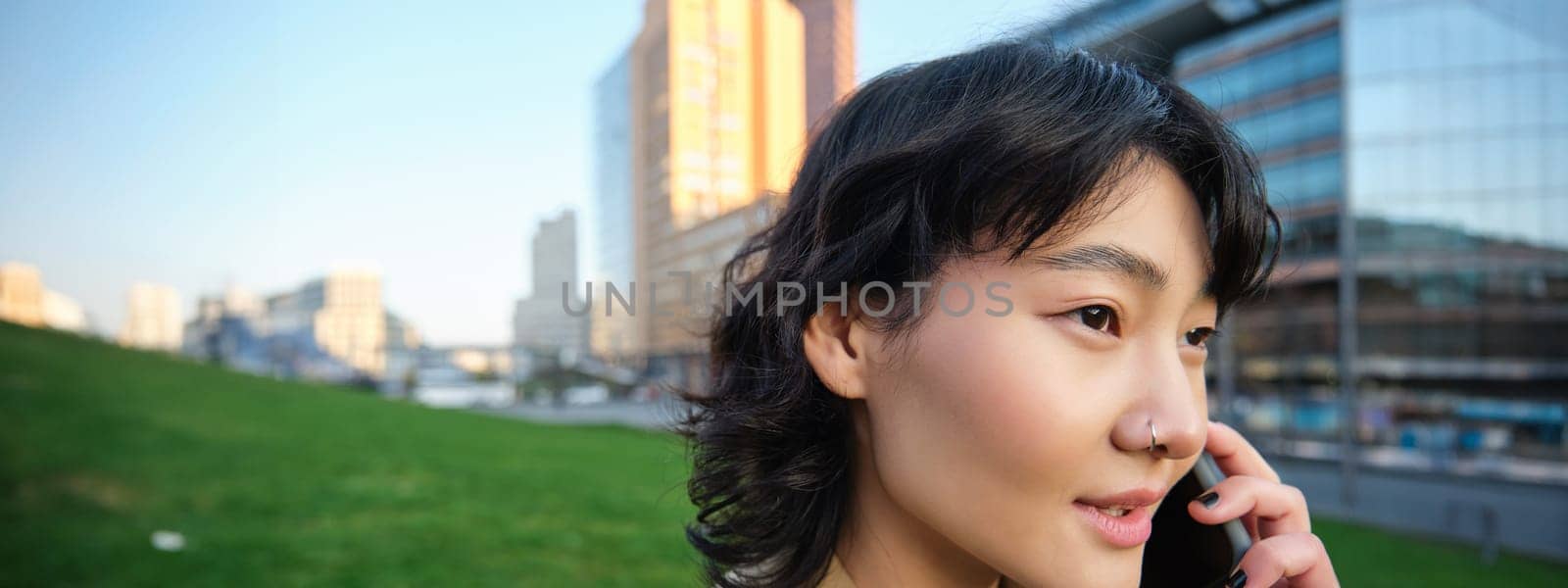 Headshot of young korean woman walks on street, talks on mobile phone, has telephone conversation, stands near campus building by Benzoix