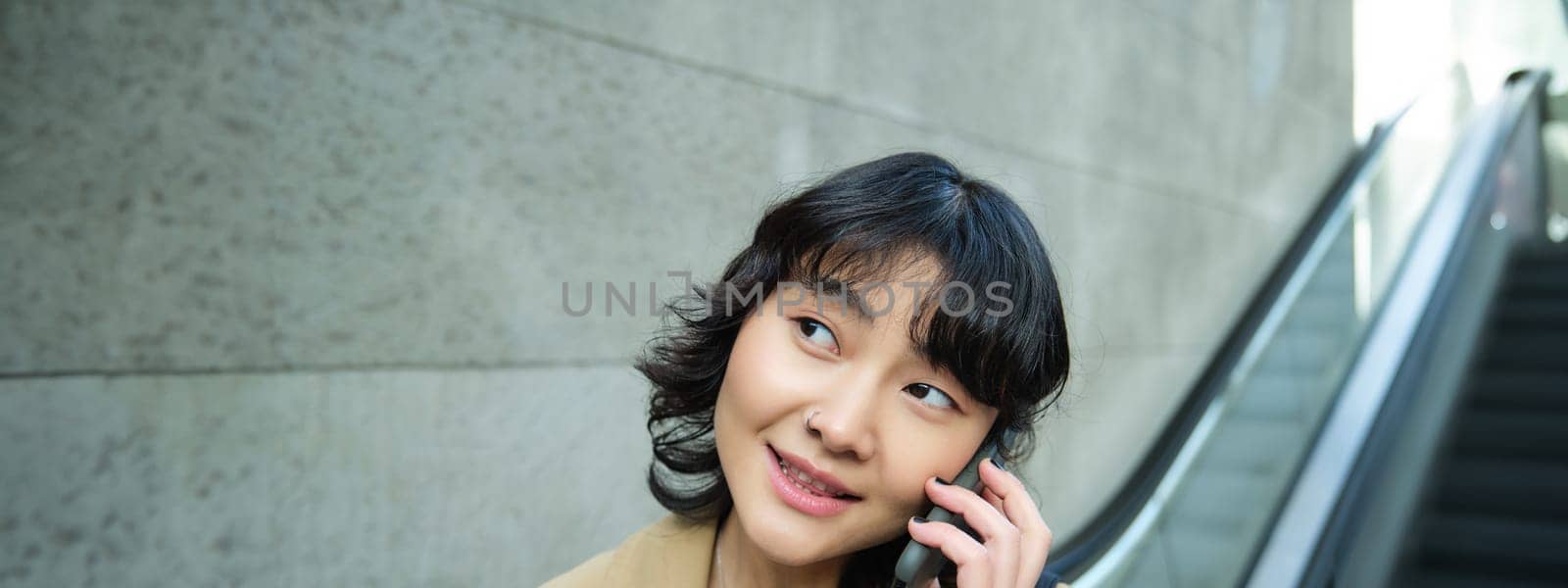 Close up portrait of smiling brunette girl, korean woman goes down escalator, makes phone call, talks to someone on telephone by Benzoix