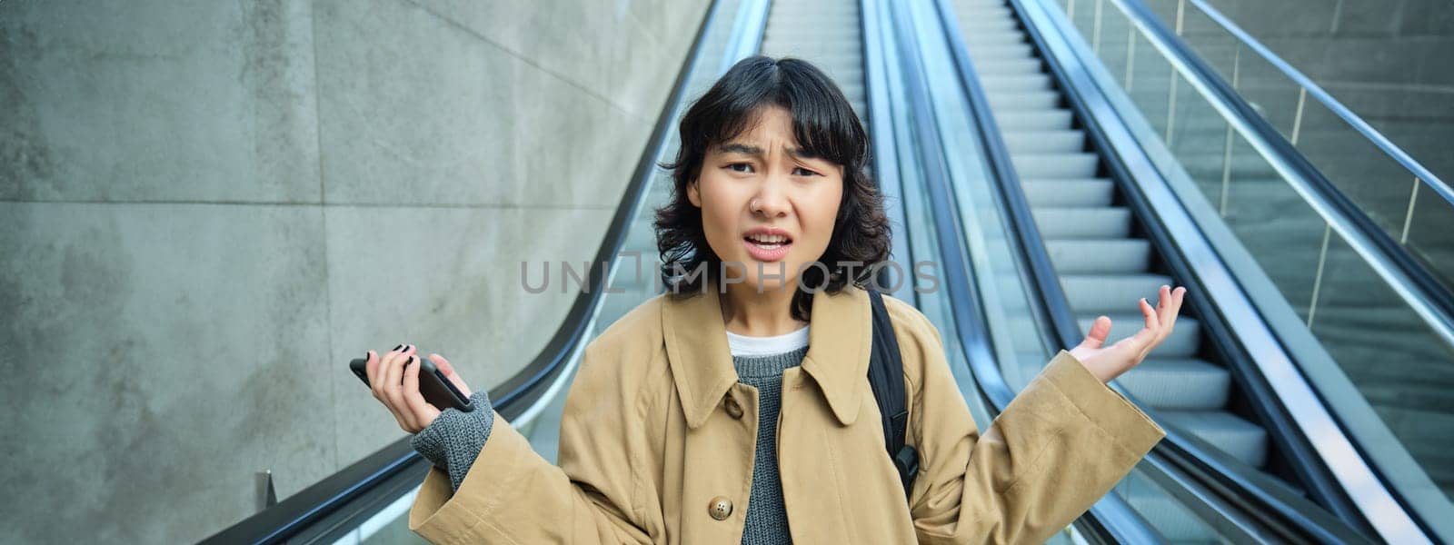 Portrait of confused asian girl doesn't know where she is, lost in unfamiliar city, goes down escalator with frustrated face, shrugging and looking at camera.