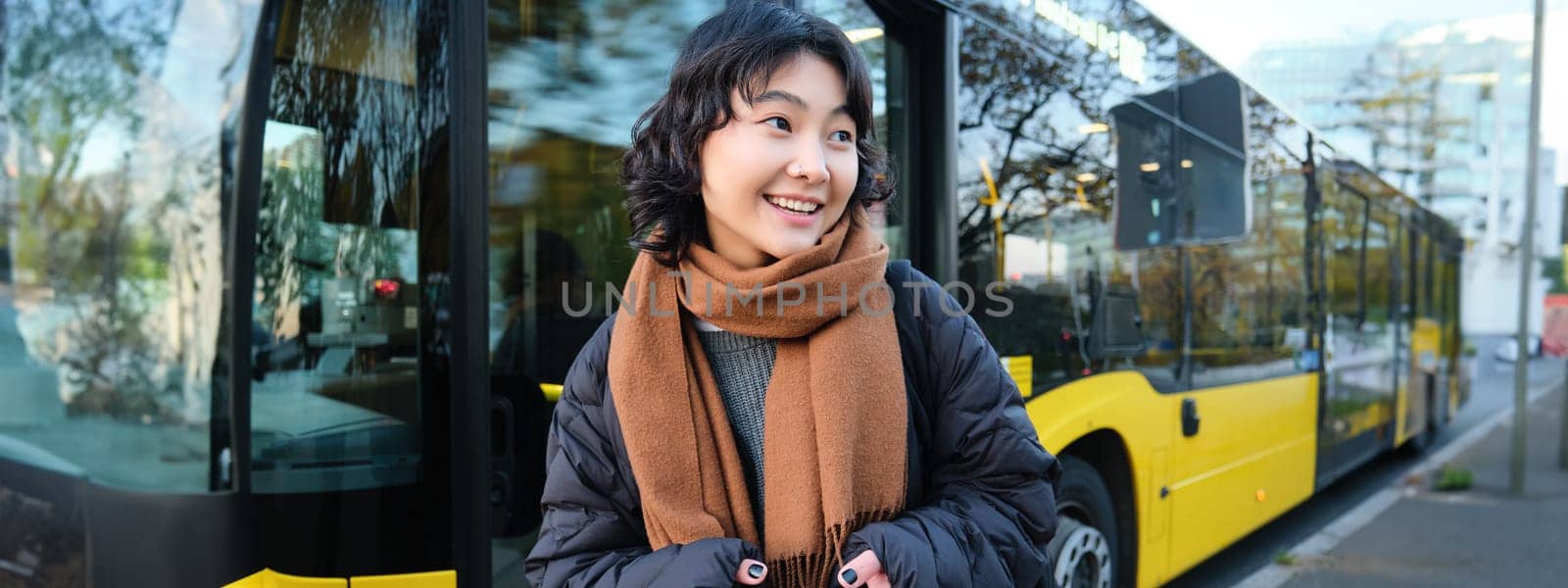 Young beautiful woman standing on bus stop, texting message on smartphone, holding mobile phone, checking her schedule, buying ticket online, wearing winter clothes by Benzoix