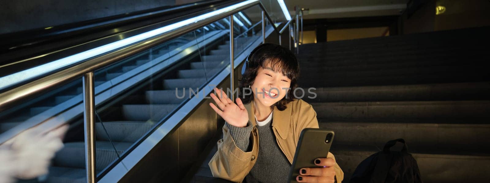 Portrait of korean girl sits on stair and waves hand at mobile phone, video chats on smartphone, greets friend on social media by Benzoix
