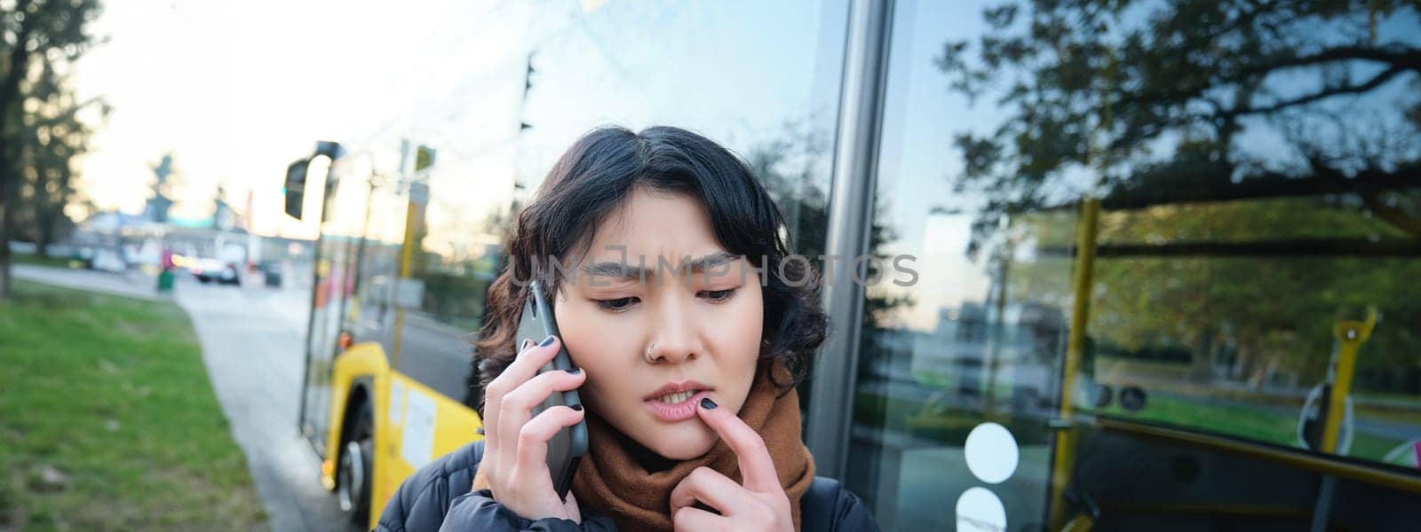 Perplexed asian girl, talks on mobile phone with concerned, thoughtful face, stands near bus stop and frowns, thinking while listening telephone conversation by Benzoix