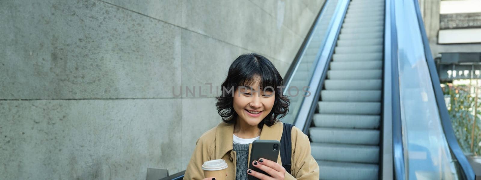 Portrait of smiling korean girl commutes, goes somewhere in city, drinks coffee to go and uses smartphone, stands on escalator by Benzoix