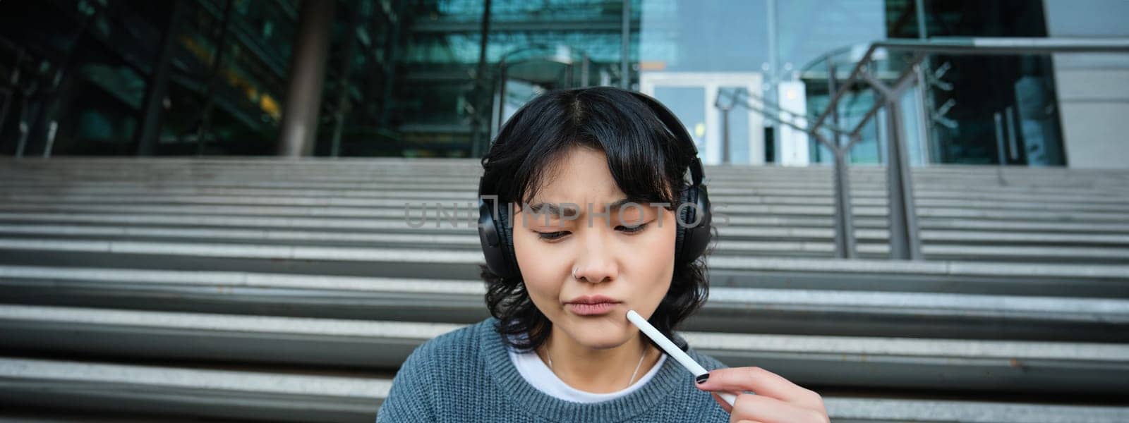 Portrait of asian girl with troubled face, draws on digital tablet with pen, looks complicated at her work, feels stuck no inspiration, sits on stairs and thinks.