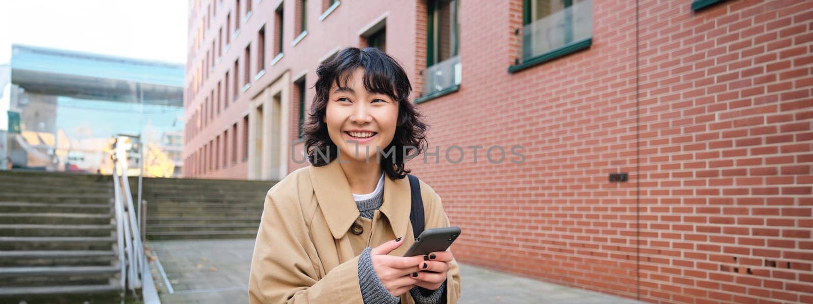 Stylish korean hipster girl, walks in city centre, sends text message, uses location map application on smartphone while walking down the street, wears trench coat by Benzoix