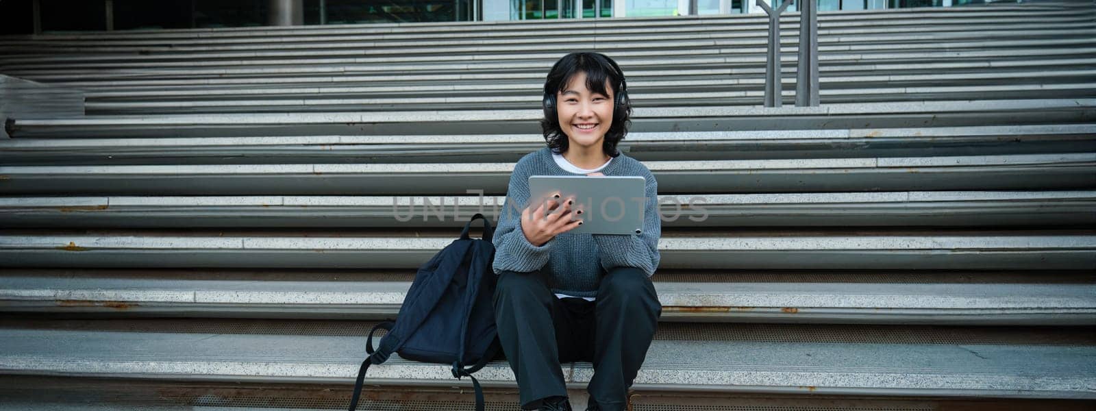 Young woman draws on her tablet, listens to music in headphones. Asian girl sits on stairs and does graphic design project, sits on street stairs.