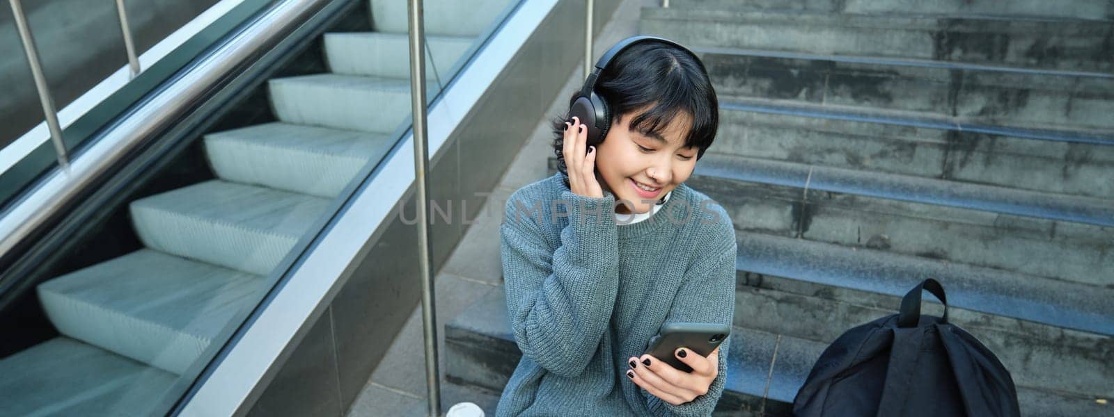 Portrait of smiling korean girl in headphones, uses smartphone and sits on stairs in mall, watches video on mobile phone by Benzoix