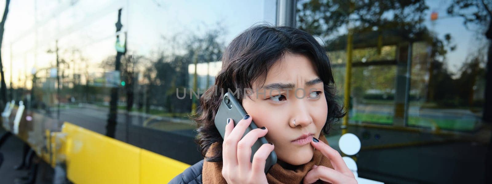 Perplexed asian girl, talks on mobile phone with concerned, thoughtful face, stands near bus stop and frowns, thinking while listening telephone conversation.