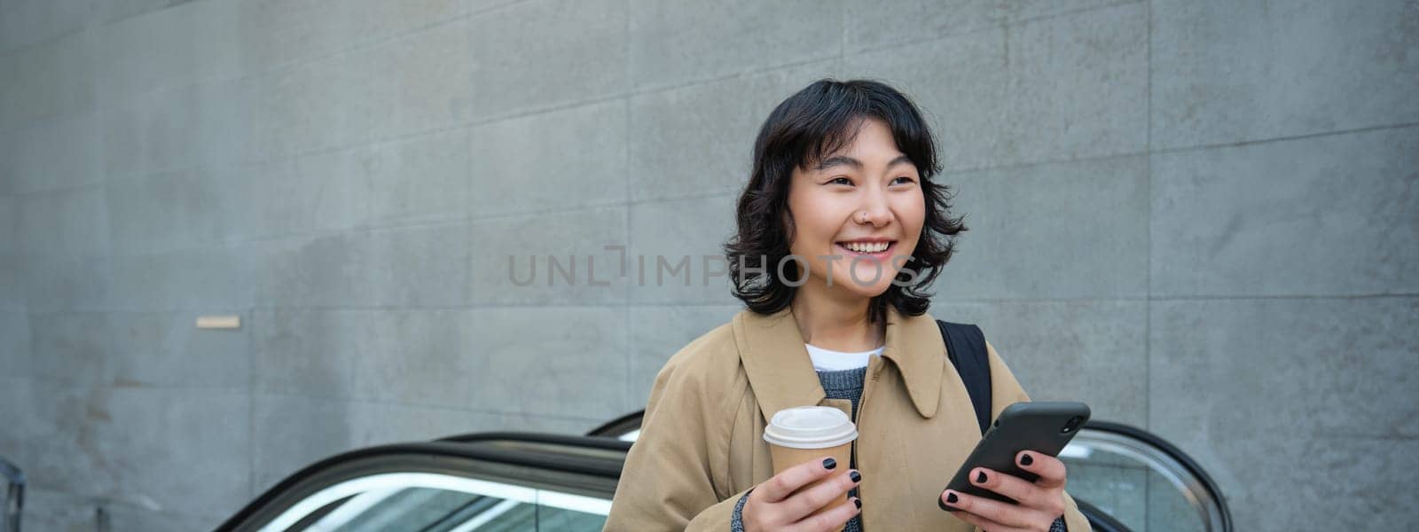 Beautiful happy asian girl, drinks coffee to go, using mobile phone while standing on escalator, walking in city centre and smiling by Benzoix