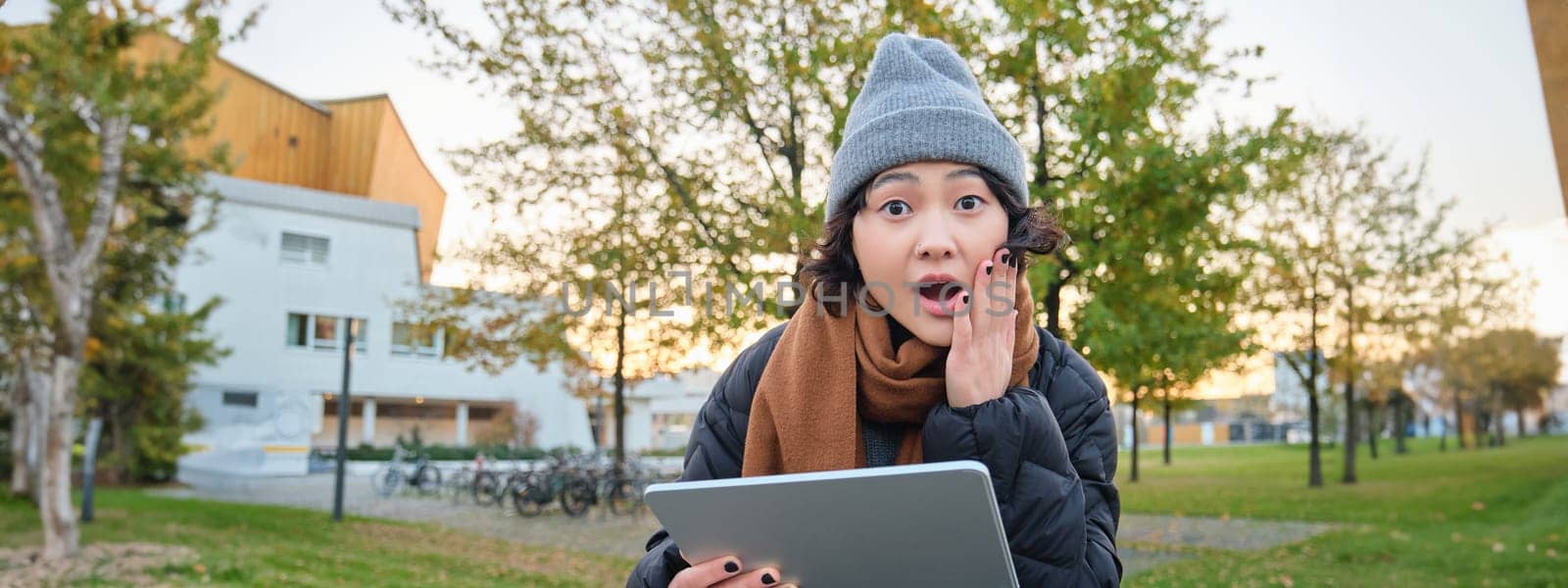 Portrait of asian girl with shocked, surprised face, holds tablet, read or watched big news, looks amazed, sits in park on bench, wears warm clothes.