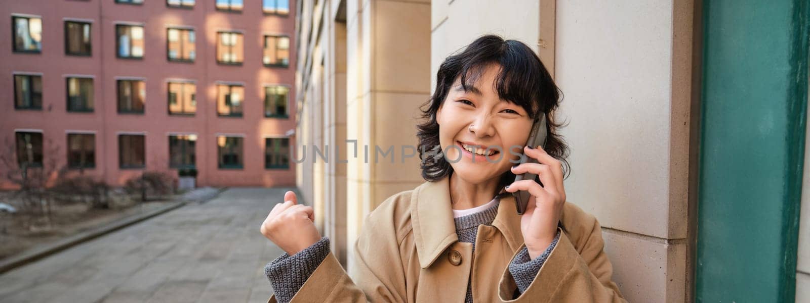 Portrait of smiling happy korean girl, stands on street, receives good news over phone, talks on smartphone and makes fist pump, celebrates, feels excitement.