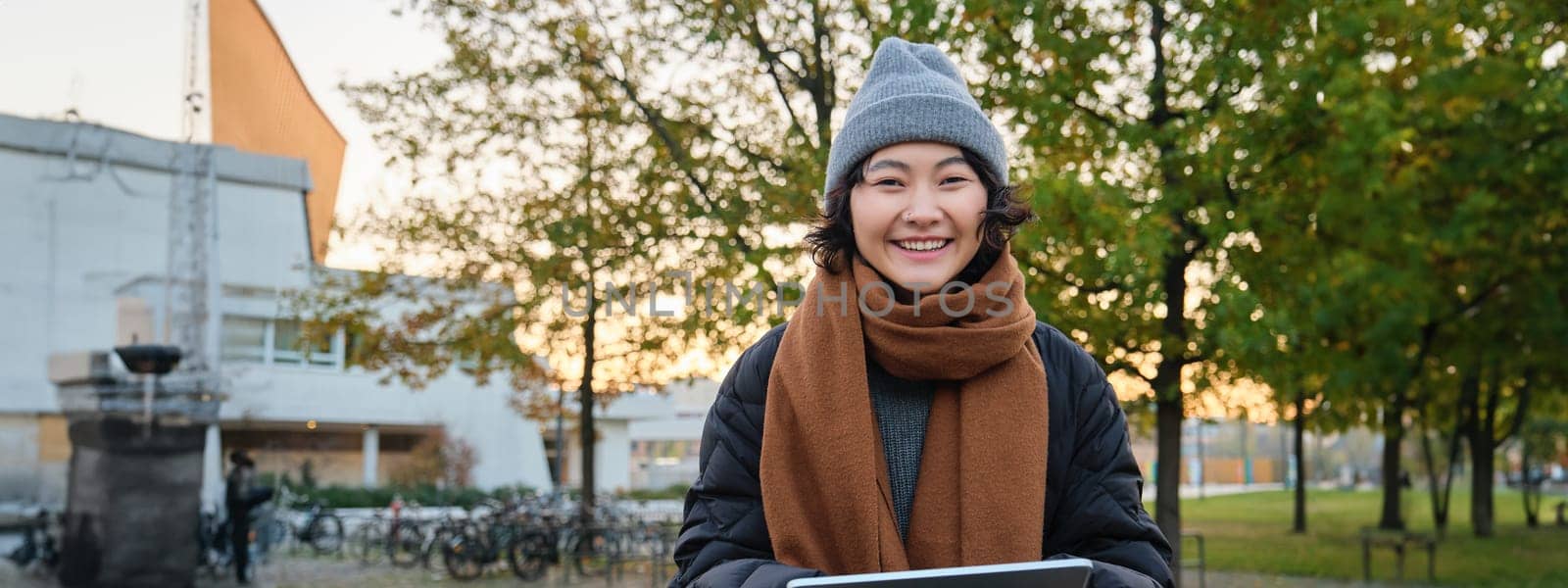 Portrait of asian girl relaxing in park, watching videos or reading on digital tablet, sitting on bench in hat and scarf on chilly day, enjoing outdoors by Benzoix