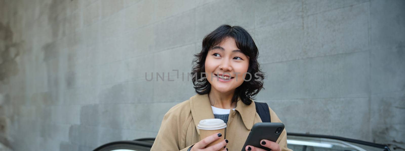 Portrait of hipster girl on escalator, drinking coffee and looking at smartphone, going to work, commute in city, smiling at camera by Benzoix