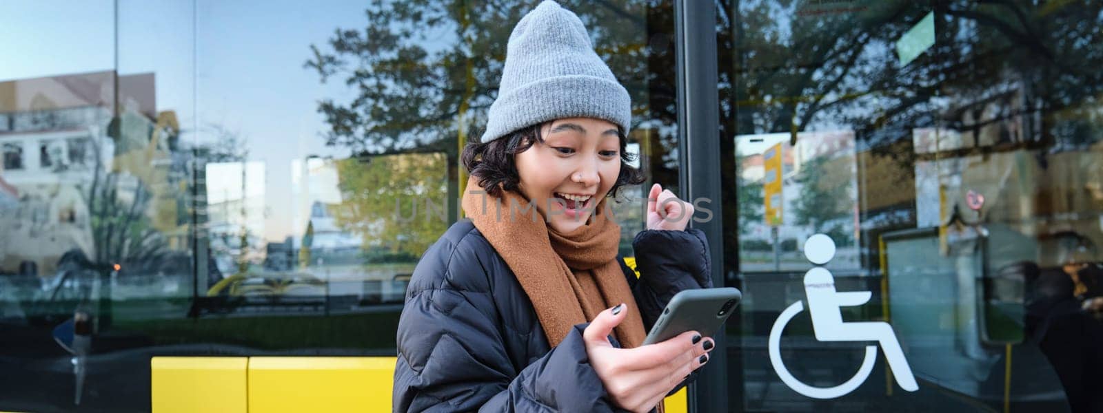 Modern people and lifestyle. Happy asian girl screams from joy, celebrates, stands near bus public transport and looks amazed.