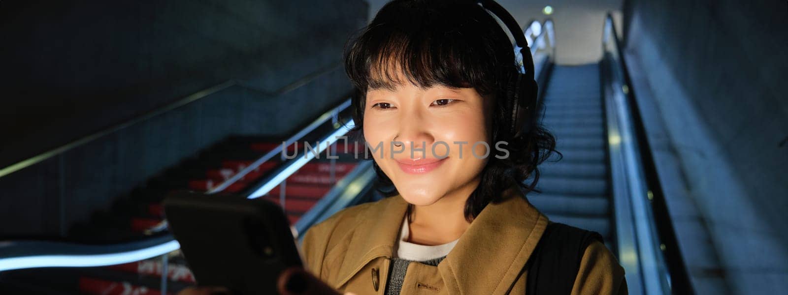 Close up of cute asian girl in headphones, picks song to listen while commuting, going down escalator to metro, smiling as reading message on mobile phone.